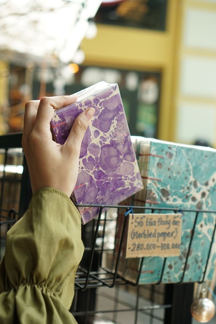 A person holds a marbled purple notebook in a stationery store setting with bright lighting.
